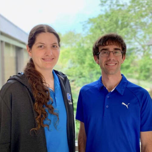 A woman and a man are standing side by side indoors, smiling at the camera with large windows and trees in the background. The woman has long curly hair, wears a blue shirt with a dark jacket, and the man wears glasses and a blue Puma polo shirt.
